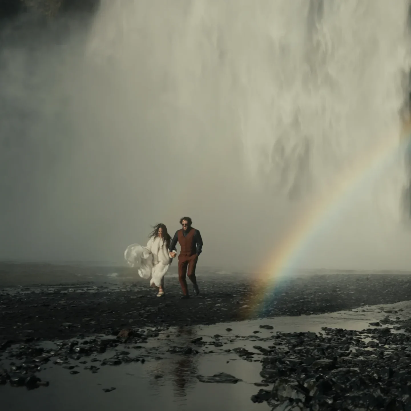 An elopement couple running in front of Skógafoss waterfall with a rainbow in the mist