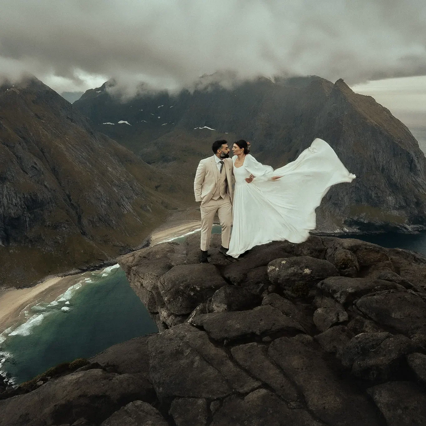 The bride and groom are standing on a rocky cliff in Norway above Kvalvika Beach, the wind lifting her dress