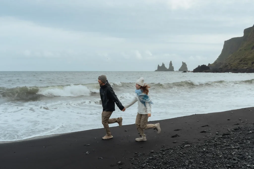Couple running hand in hand along a black sand beach in Iceland, with waves, rugged cliffs, and sea stacks in the background.