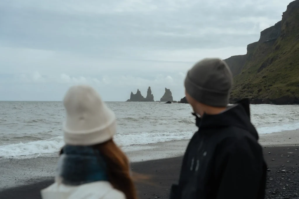 Couple looking out at Reynisdrangar sea stacks from a black sand beach in Iceland.