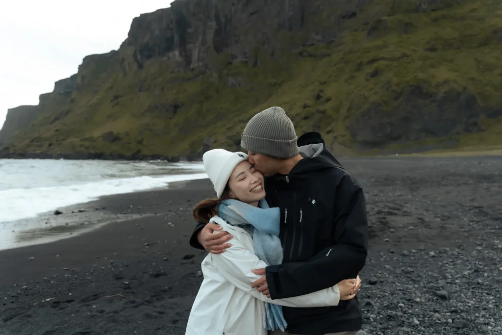 Couple embracing and smiling on a black sand beach in Iceland, with dramatic moss-covered cliffs and ocean waves in the background.