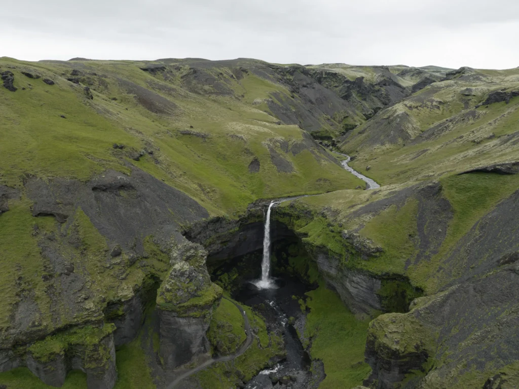 Aerial view of Kvernufoss waterfall in Iceland, surrounded by lush green moss-covered cliffs, volcanic rock formations, and a winding river.