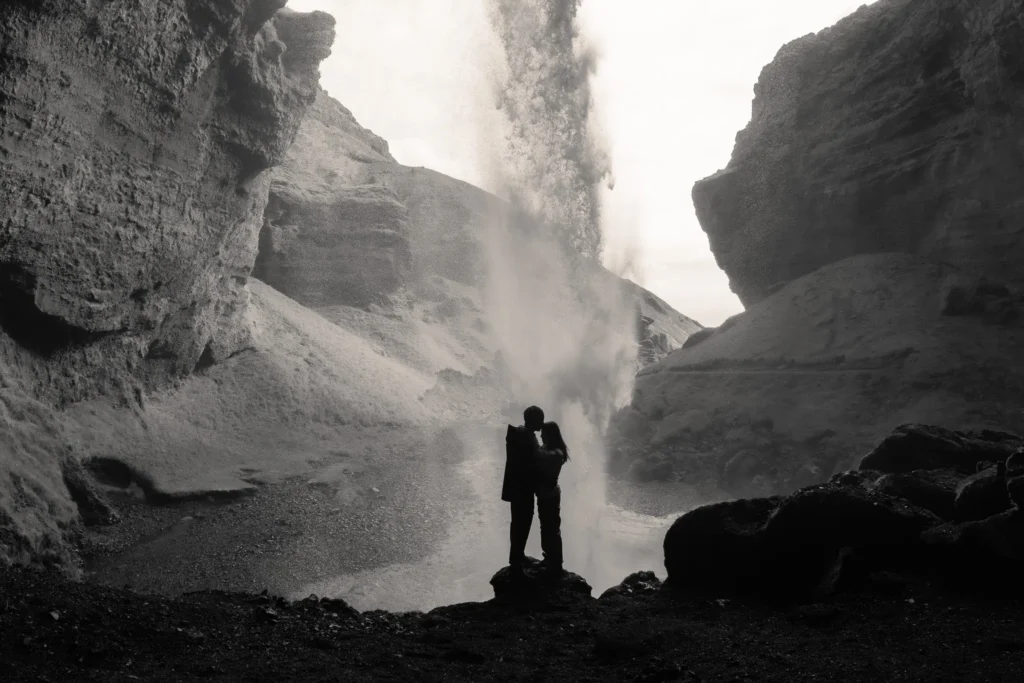 Couple embracing in front of Kvernufoss waterfall in Iceland, surrounded by dramatic volcanic cliffs and mist in a black-and-white photograph.
