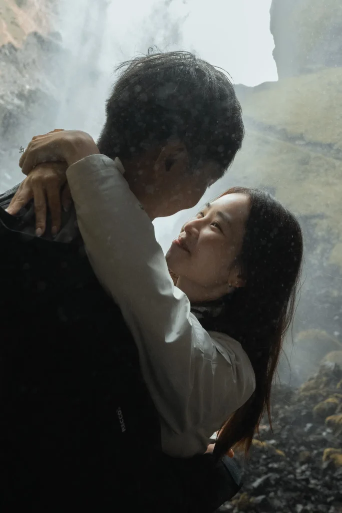 Couple embracing and sharing an intimate moment in front of Kvernufoss waterfall in Iceland, surrounded by mist and dramatic volcanic cliffs.