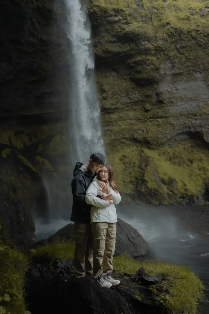 Couple embracing in front of Kvernufoss waterfall in Iceland, surrounded by moss-covered cliffs and misty volcanic landscape.