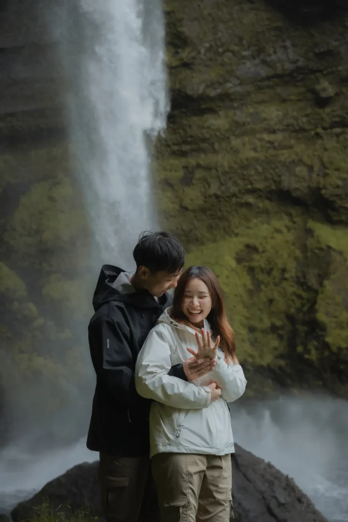 Couple embracing in front of Kvernufoss waterfall in Iceland, surrounded by moss-covered cliffs and misty volcanic landscape.