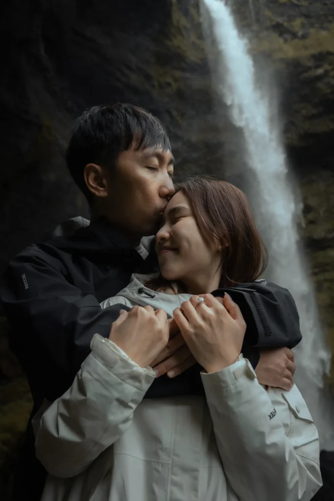 Couple embracing and sharing a tender moment in front of Kvernufoss waterfall in Iceland, surrounded by mist and moss-covered cliffs.