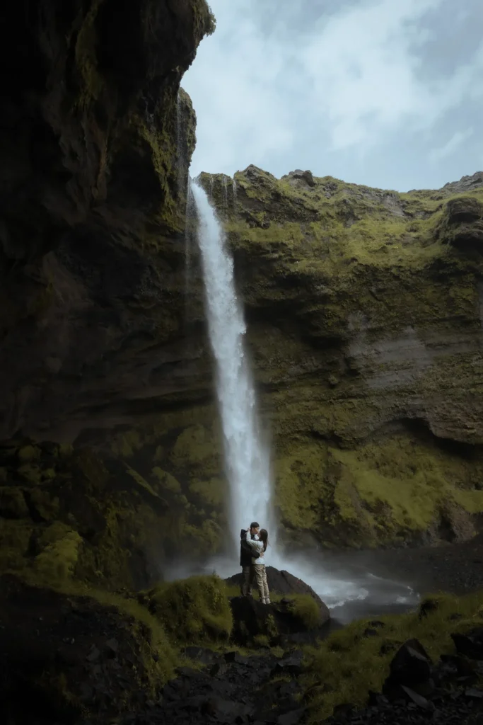 Couple embracing in front of Kvernufoss waterfall in Iceland, surrounded by dramatic moss-covered cliffs and volcanic rock formations.