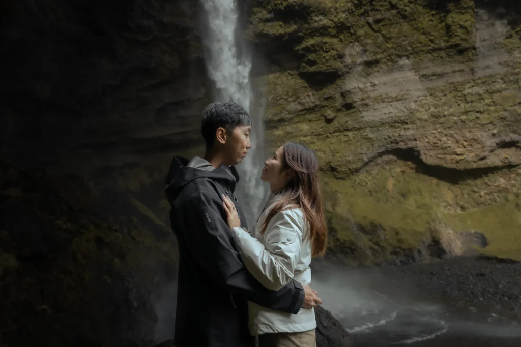Couple standing together in front of Kvernufoss waterfall in Iceland, surrounded by moss-covered volcanic cliffs and mist.