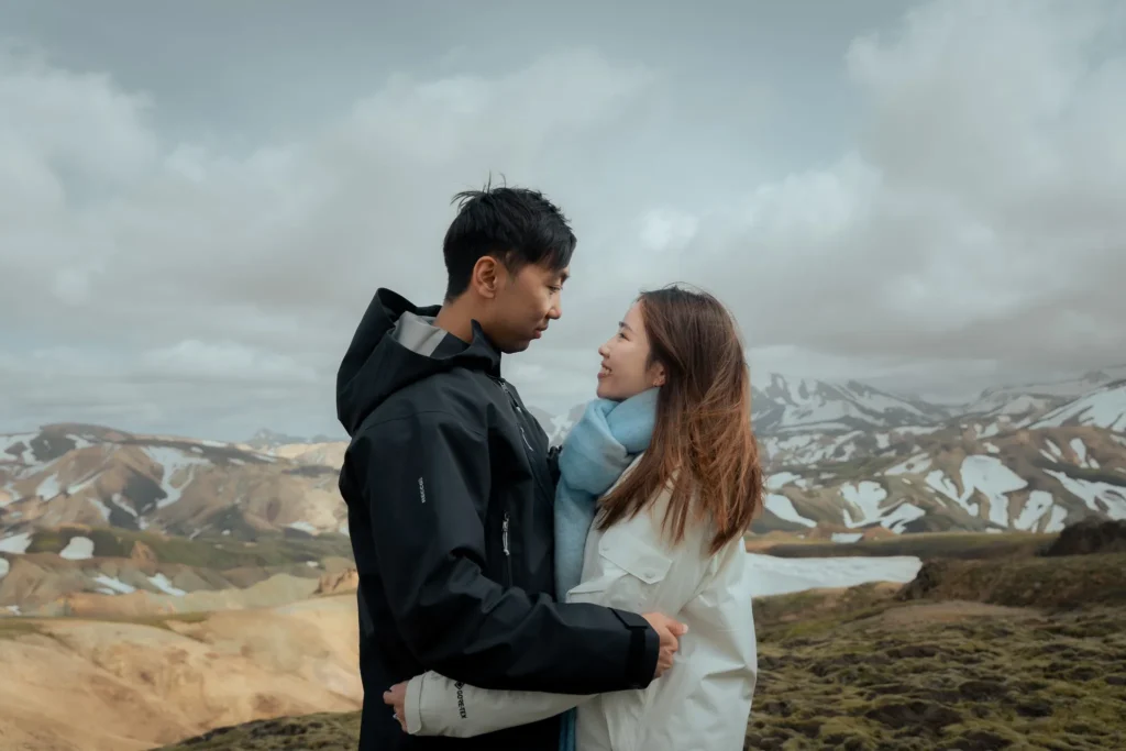 Engagement couple embracing and looking at each other in Landmannalaugar, Iceland, surrounded by colorful volcanic mountains and snow-covered peaks.