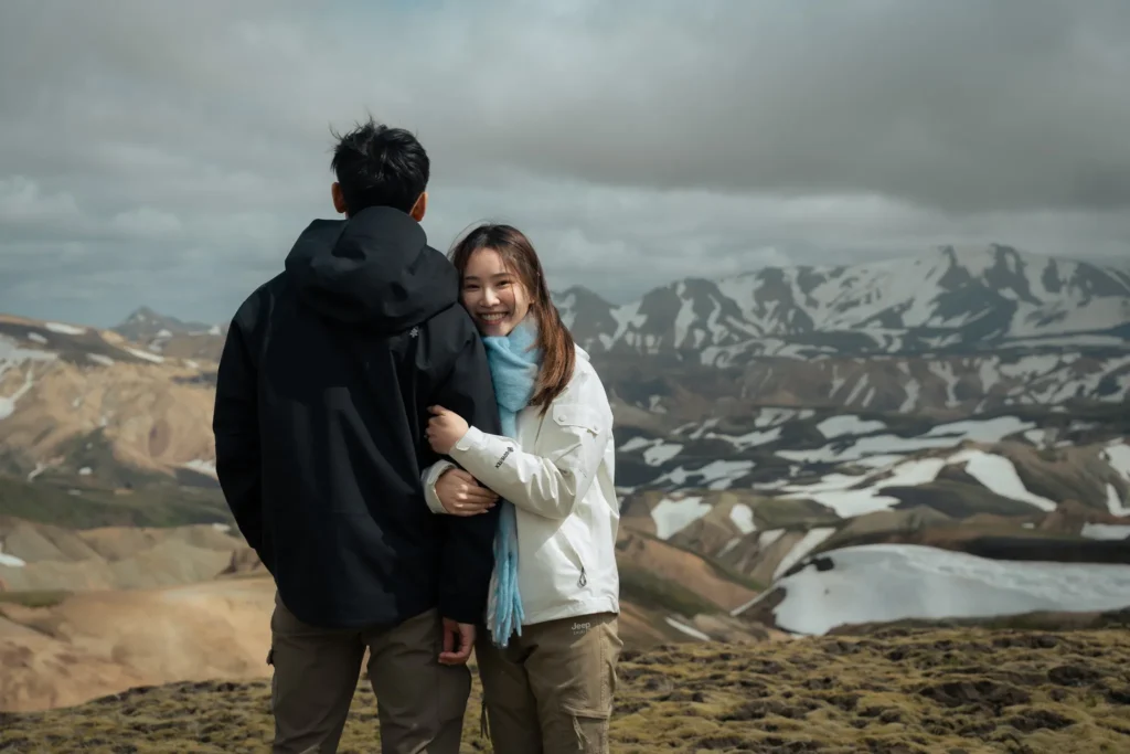 Engagement couple embracing in Landmannalaugar, Iceland, with colorful volcanic mountains, snow-covered peaks, and a glacial lake in the background.