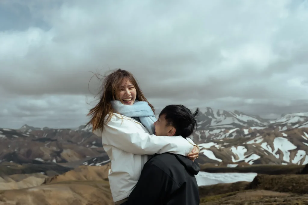 **Alt description:** Engaged couple sharing a joyful moment in Landmannalaugar, Iceland, surrounded by colorful mountains, snow-covered peaks, and dramatic clouds.