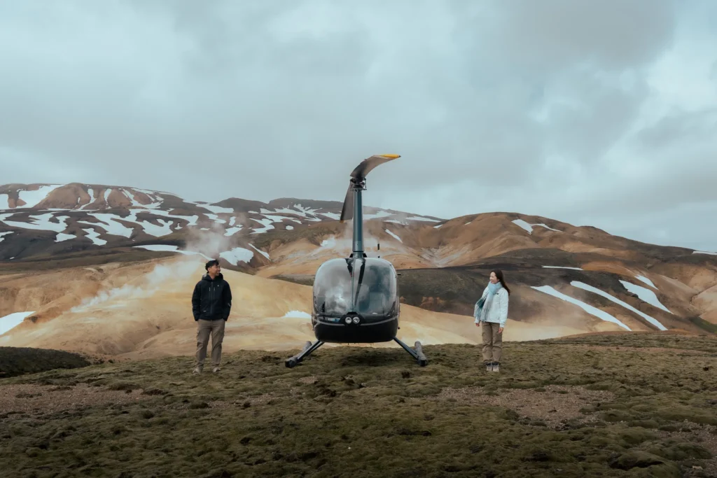 Engaged couple standing beside a helicopter in Landmannalaugar, Iceland, surrounded by colorful volcanic mountains, snow patches, and geothermal steam.