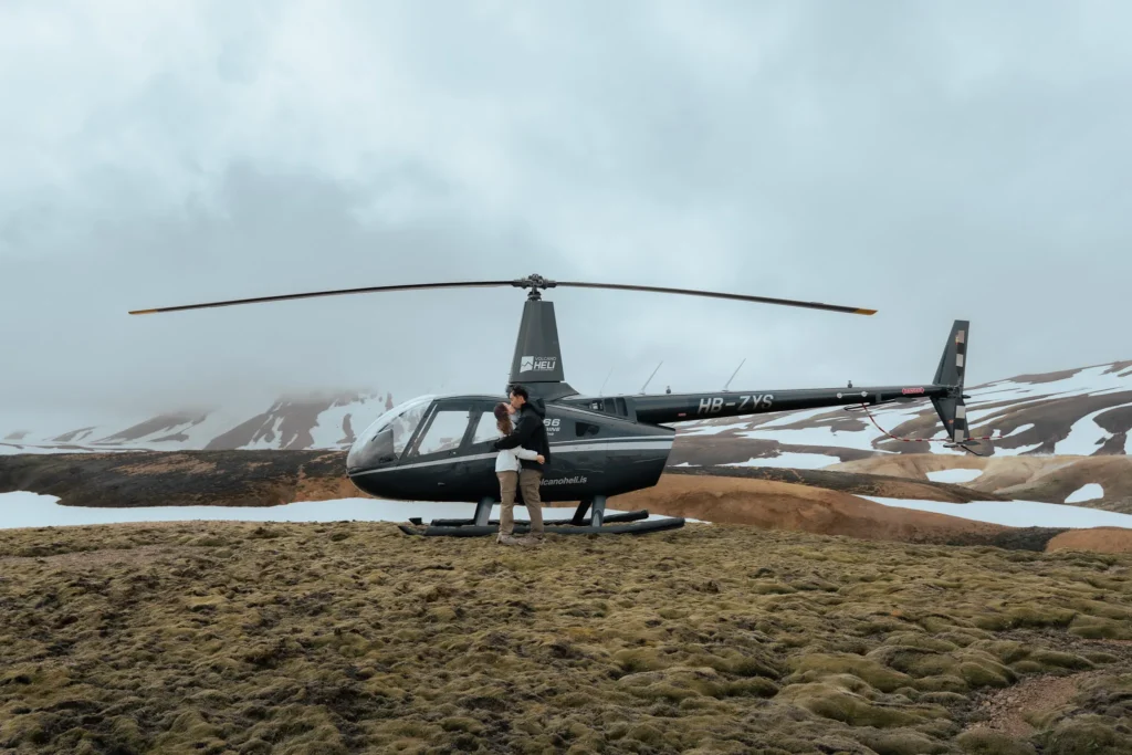 Engaged couple embracing beside a helicopter in the Icelandic highlands of Landmannalaugar, surrounded by moss-covered terrain and snow-capped mountains.
