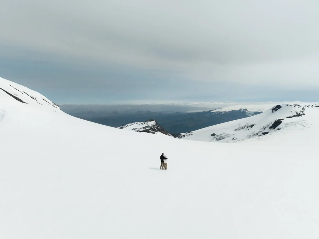 Couple standing together on a vast snow-covered Icelandic mountain landscape, surrounded by glaciers, snowy peaks, and distant mountains under a cloudy sky.