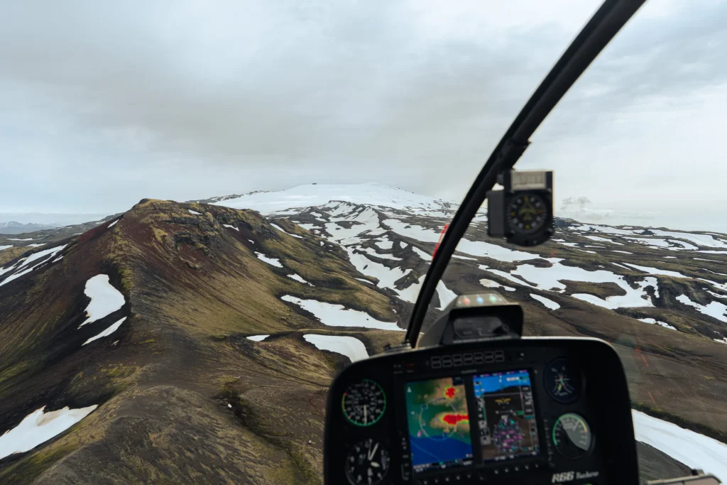 View from inside a helicopter flying over Icelandic volcanic mountains with patches of snow and a rugged landscape.