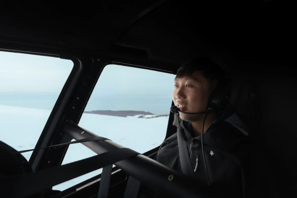 A man wearing a headset inside a helicopter, looking out over a snow-covered Icelandic landscape.