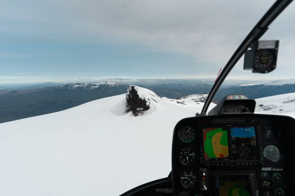 View from inside a helicopter flying over a snow-covered Icelandic glacier and surrounding mountain landscape.
