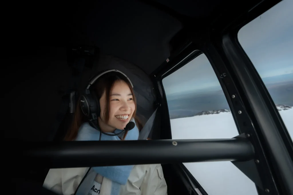 A woman wearing a headset and smiling inside a helicopter, with a snowy Icelandic landscape visible through the window.