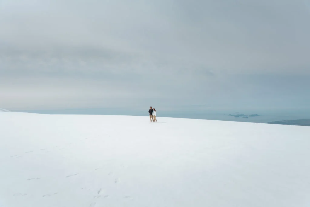 A couple walking across a vast snowy glacier in Iceland, surrounded by an expansive white landscape and distant ocean views.