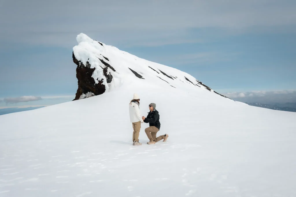 A man proposing to his partner on a snowy glacier in Iceland, with a dramatic snow-covered mountain behind them.
