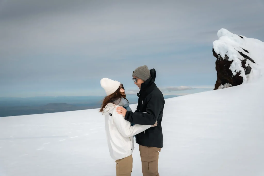 A couple embracing on a snowy glacier in Iceland, with a dramatic snow-covered mountain and distant landscape in the background.
