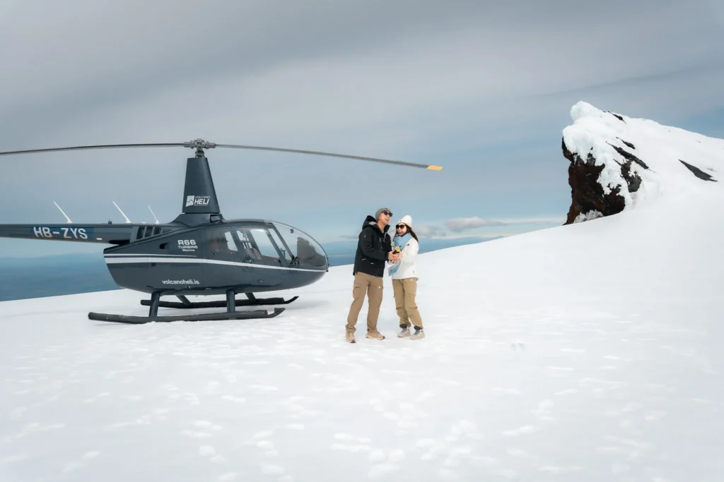 A couple celebrating beside a helicopter on a snowy glacier in Iceland, with a dramatic snow-covered mountain in the background.