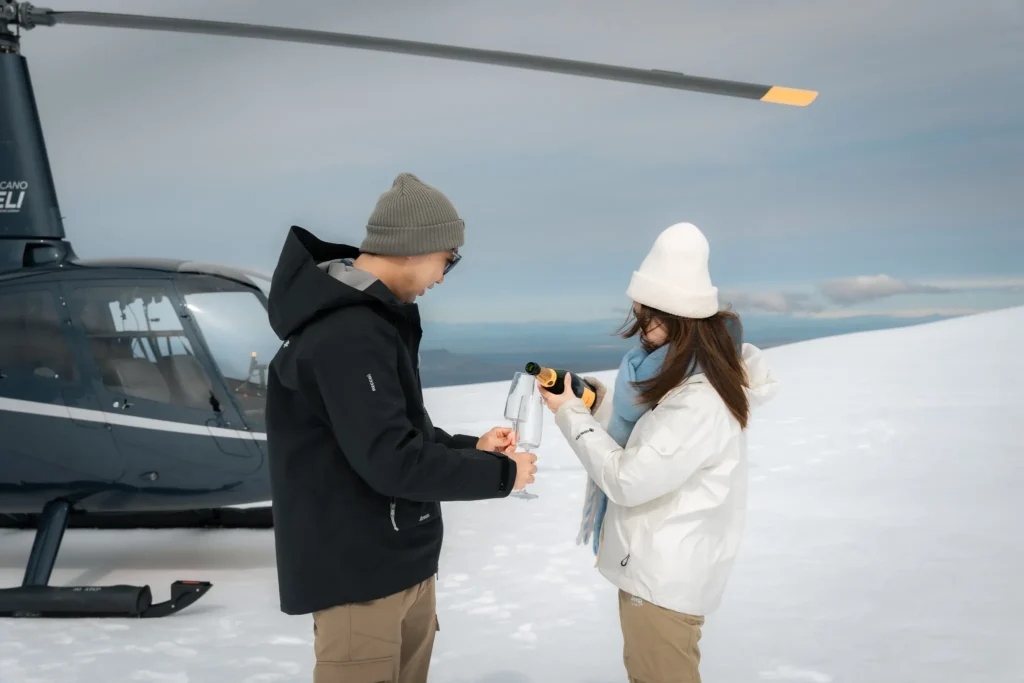 A couple pouring champagne into glasses beside a helicopter on a snowy glacier in Iceland.