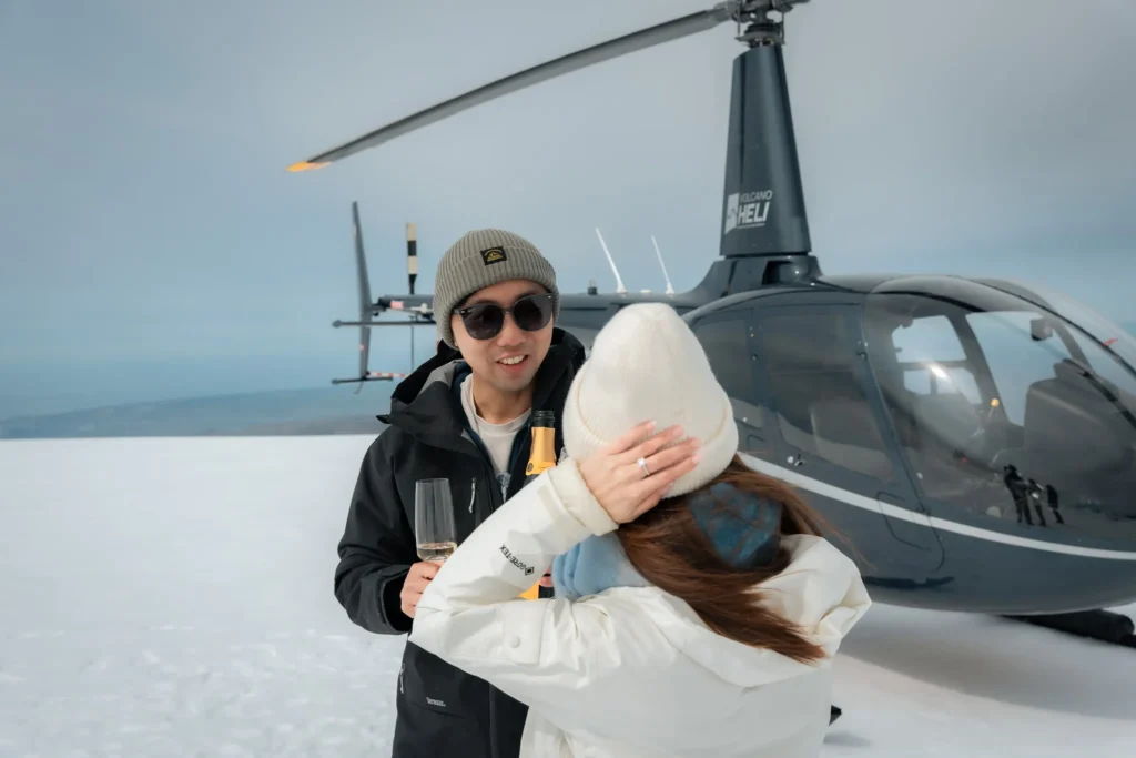 A couple celebrating on a snowy glacier beside a helicopter, with champagne and an engagement ring visible.