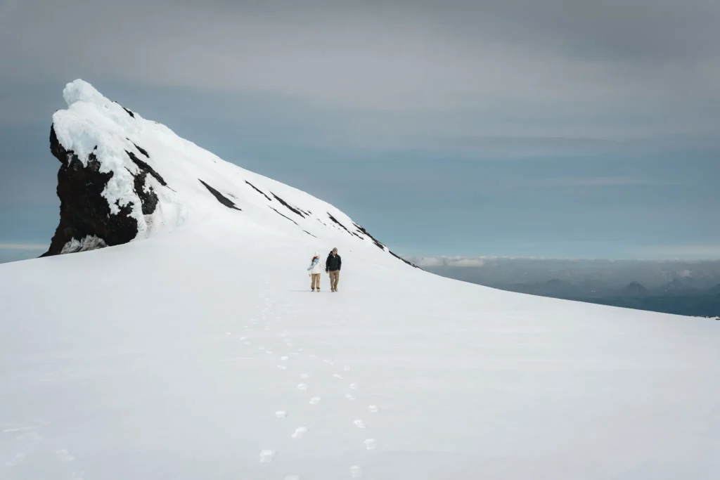 Two people walking across a snowy glacier beneath a dramatic snow-covered mountain in Iceland, with distant mountains visible on the horizon.