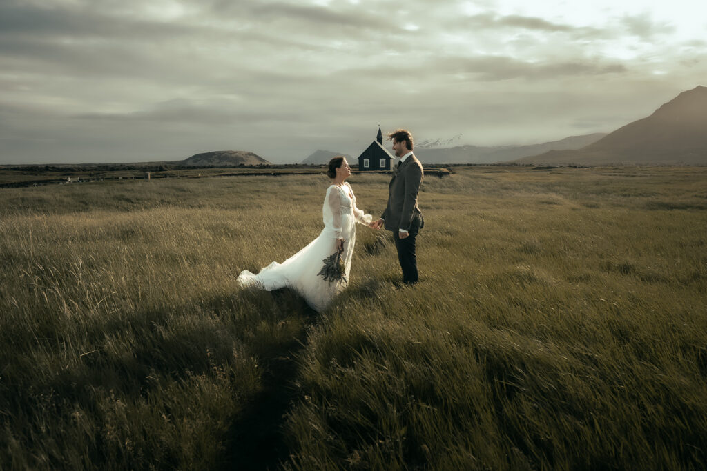 A bride and groom holding hands in a grassy landscape with a black church Budakirkja in the background in Iceland