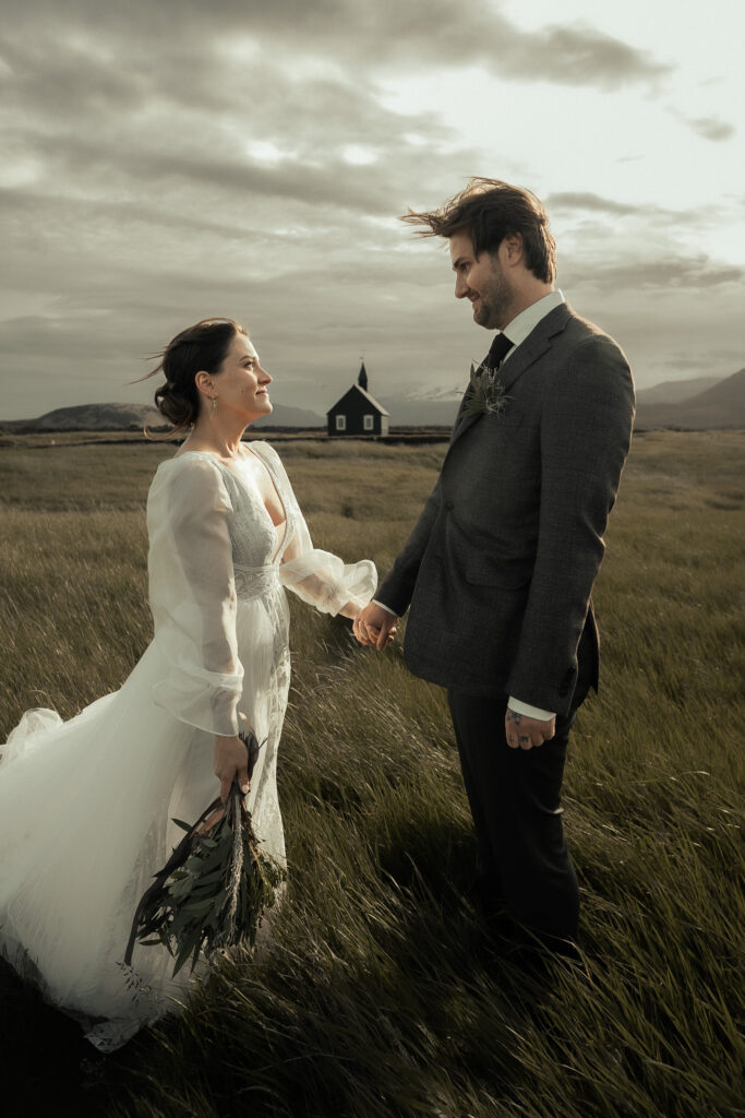 A bride and groom holding hands in a grassy landscape with a black church Budakirkja in the background in Iceland