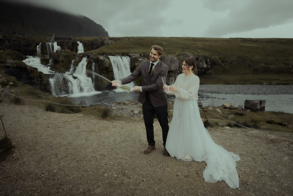 A bride and groom opening a bottle of champagne near waterfalls at Kirkjufell in Iceland
