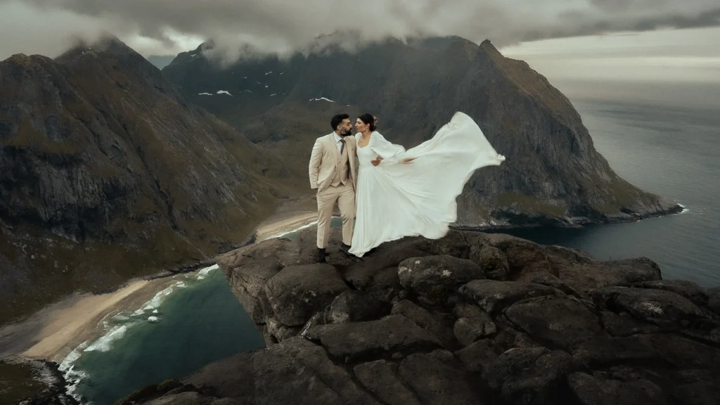 The bride and groom are standing on a rocky cliff in Norway above Kvalvika Beach, the wind lifting her dress.