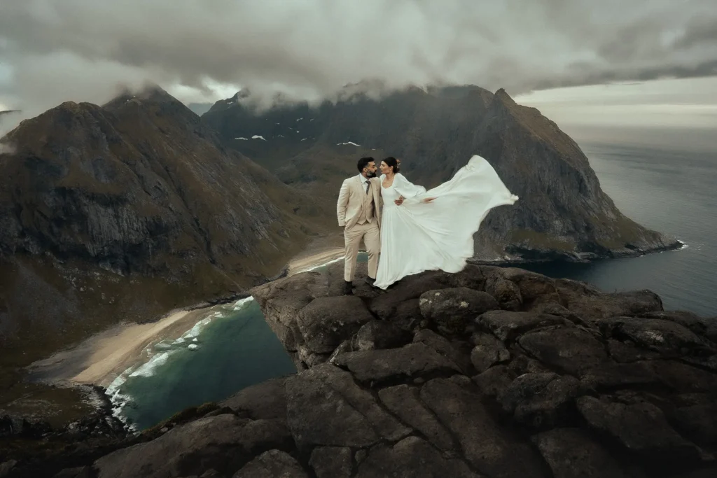 The bride and groom are standing on a rocky cliff in Norway above Kvalvika Beach, the wind lifting her dress