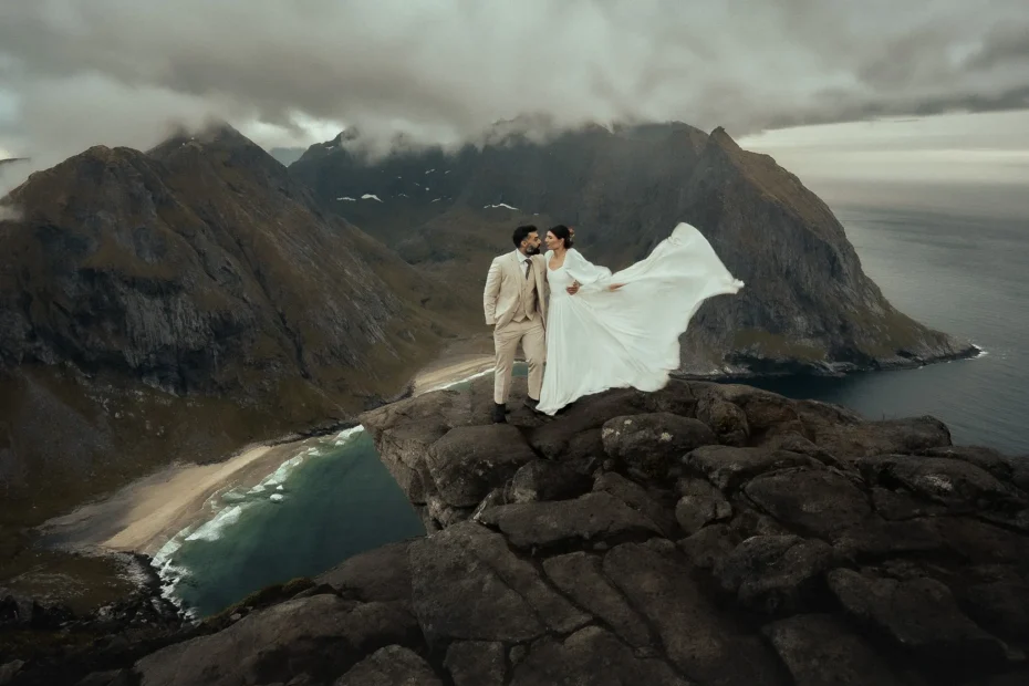 The bride and groom are standing on a rocky cliff in Norway above Kvalvika Beach, the wind lifting her dress