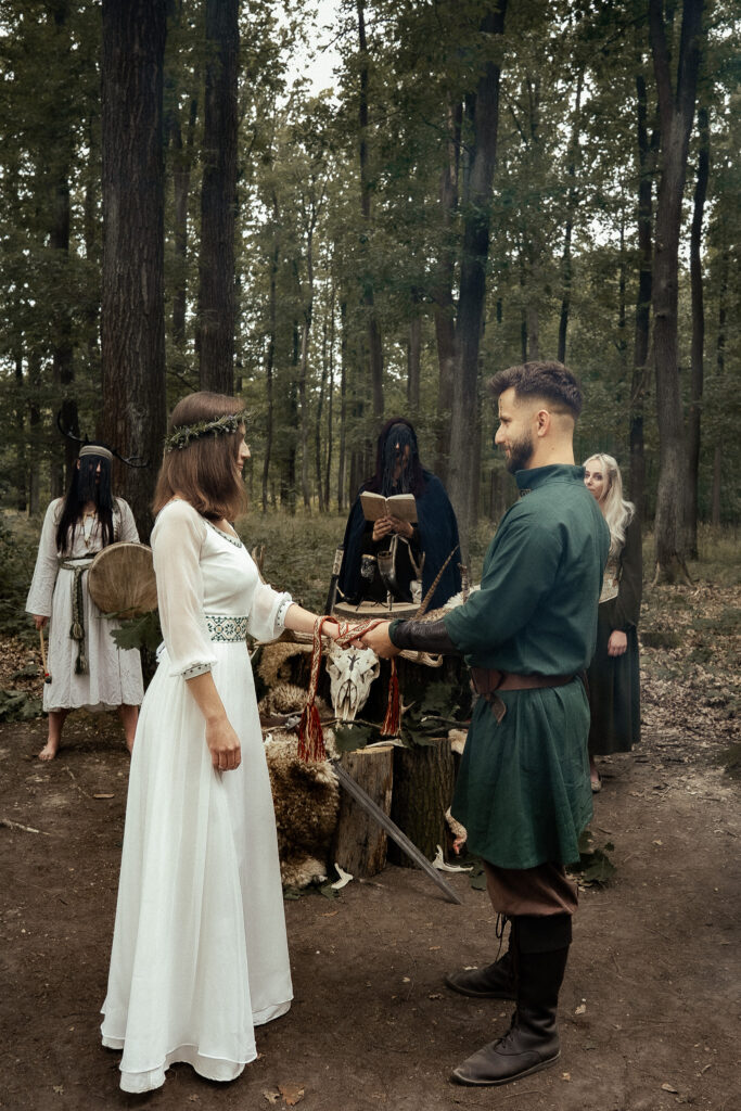 Pagan wedding ceremony in the forest with bride and groom holding hands tied by a red ribbon