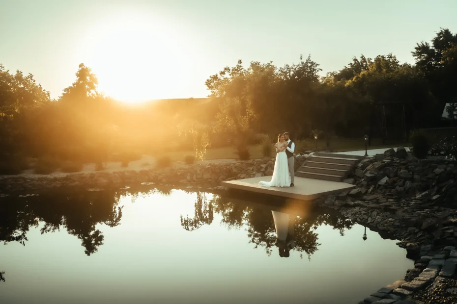 Pár stojí na dřevěném molu u rybníka při západu slunce na svatebním místě Nový Dvůr