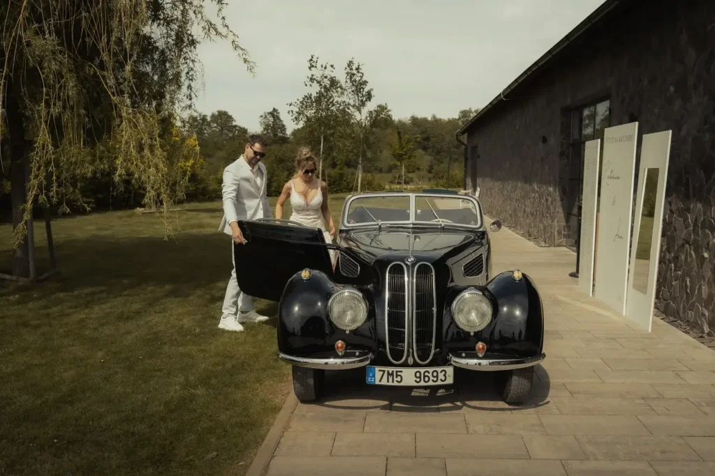Nevěsta a ženich nastupují do veteránského auta ve svatebním areálu Stodola Prost